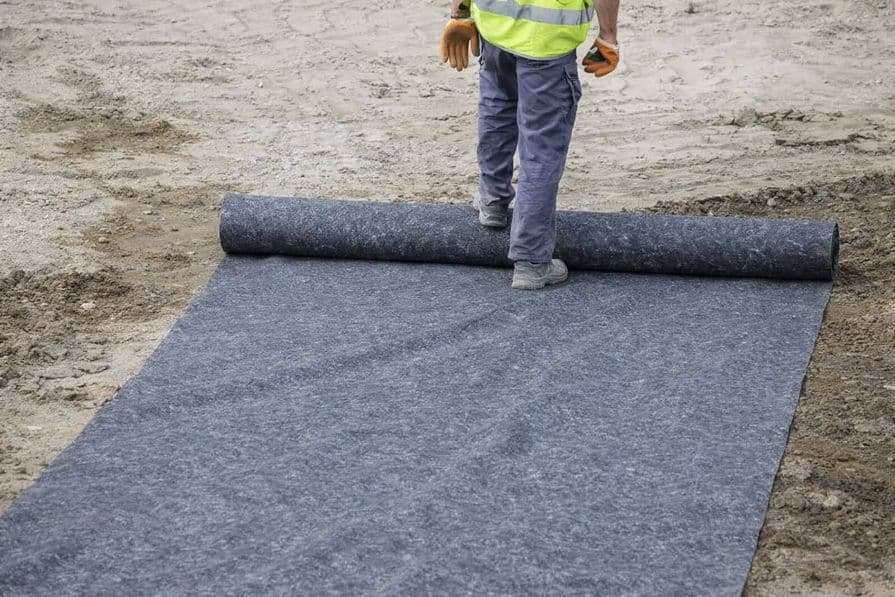 Pokládka netkanej geotextílie na upravený podklad. Pracovník v reflexnej veste rozvinie rolku geotextílie na stavenisku